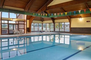 Ein Innenpool mit klarem, blauem Wasser, Holzbalken an der Decke und Blick durch große Fenster nach draußen.