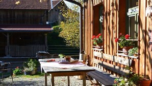 Une terrasse extérieure en bois, avec une table et un banc, entourée de plantes en pot et d’un bâtiment au style rustique.