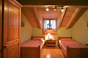 Ein Zimmer mit zwei Holzbetten, einem mit Vorhängen versehenen Fenster, einem Holzschrank und einer Holzbalkendecke.