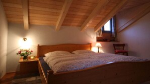 Une chambre avec un plafond en bois, un lit recouvert d’une couvre-lit à motifs floraux, une table de chevet avec une lampe et une fenêtre donnant sur un beau paysage.