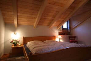 Ein Schlafzimmer mit Holzbalkendecke, einem Bett mit floralem Überwurf, einem Nachttisch mit Lampe und einem Fenster mit Ausblick.