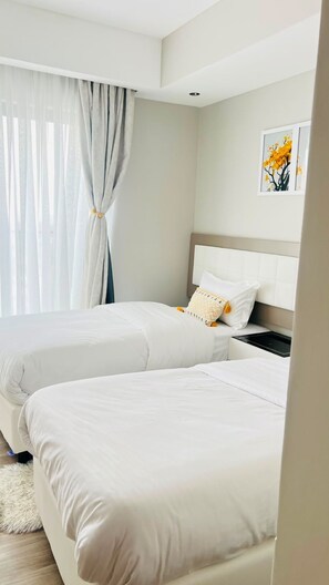 A hotel room with two beds, a large window with sheer curtains, a white headboard, and a framed picture on the wall.