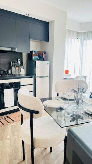 A modern kitchen with dark cabinets, a microwave, and a refrigerator. There is a dining area with a glass table set for two, and a chair.