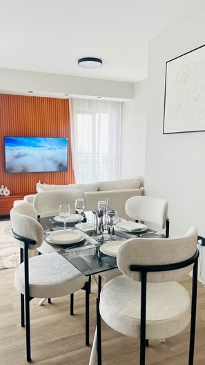 A modern dining area with a glass table, white chairs, and a flat-screen TV mounted on the wall.