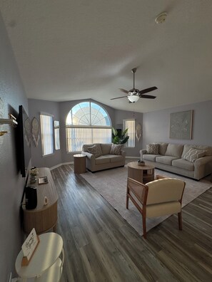 A spacious living room with a large arched window, a ceiling fan, and a flat-screen TV mounted on the wall. - Family-Friendly Kissimmee Stay Near Disney (Kissimmee)