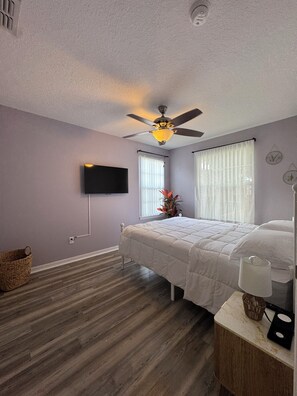 Un dormitorio con cama, ventilador de techo, televisor y ventana con cortinas.