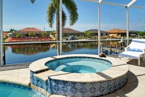 Un jacuzzi con borde de azulejos, una piscina, sillas de descanso y vistas a un edificio al otro lado del agua.