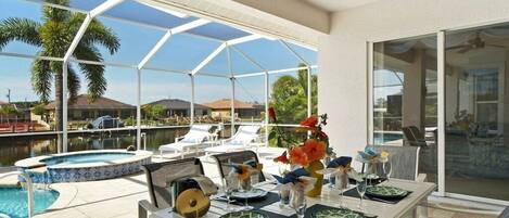 Comedor con una mesa preparada para cuatro, con vista a una piscina y un paisaje de palmeras y agua.