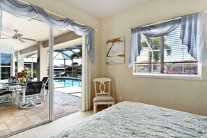 Un dormitorio con cama, una silla, ventana con cortinas, un cuadro y vista a una piscina y zona exterior.