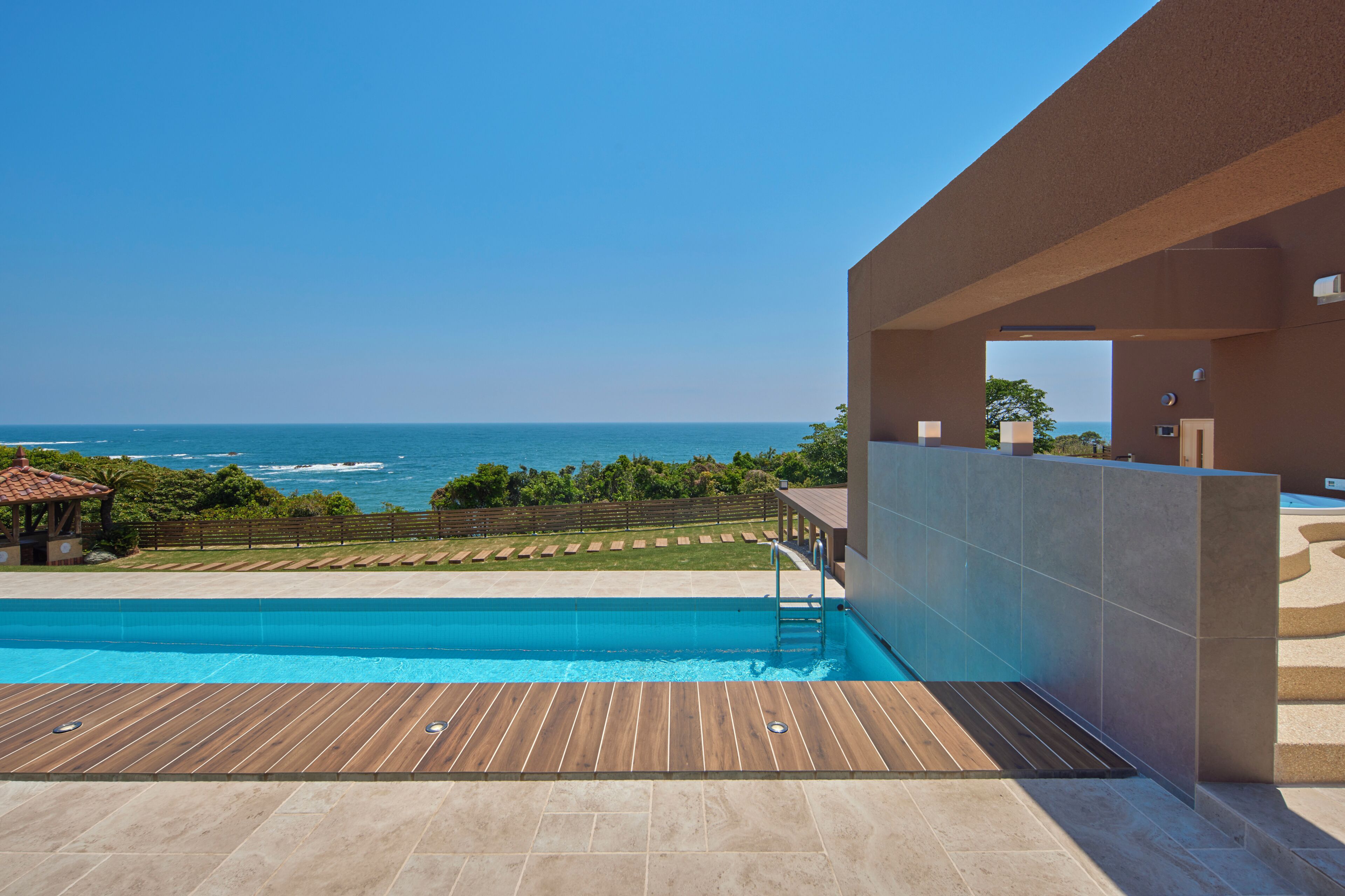 A modern pool area with a view of the ocean, wooden deck, and a covered seating area.