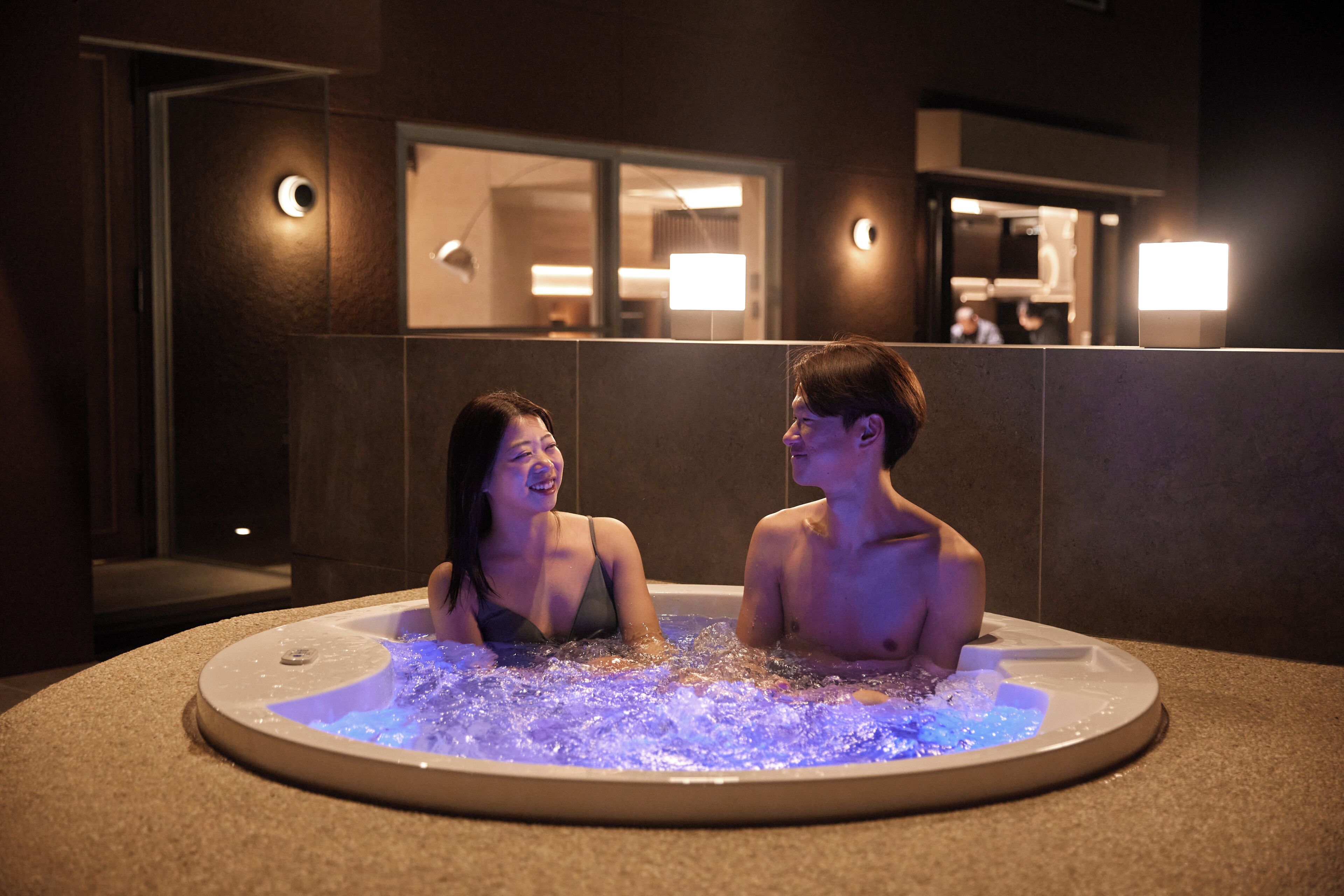 Two people in a heated, circular jacuzzi with a modern design, surrounded by a tiled wall and a doorway leading to a room with a window.