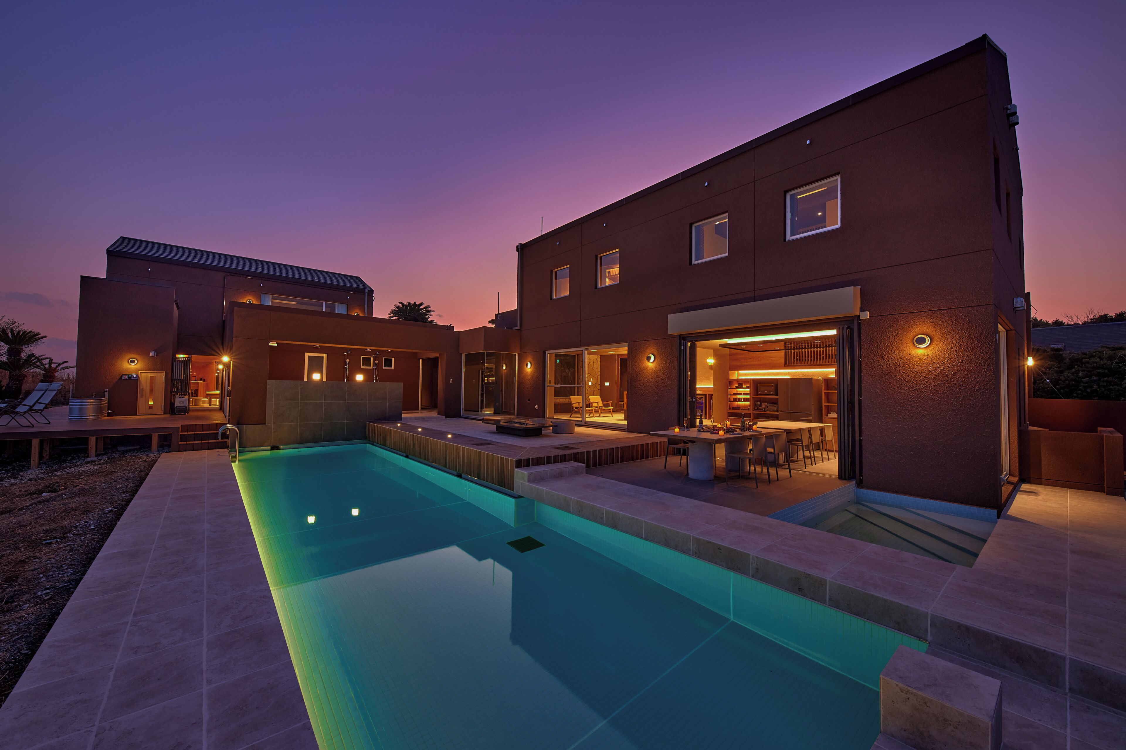A modern house with a swimming pool, outdoor seating, and a well-lit interior.