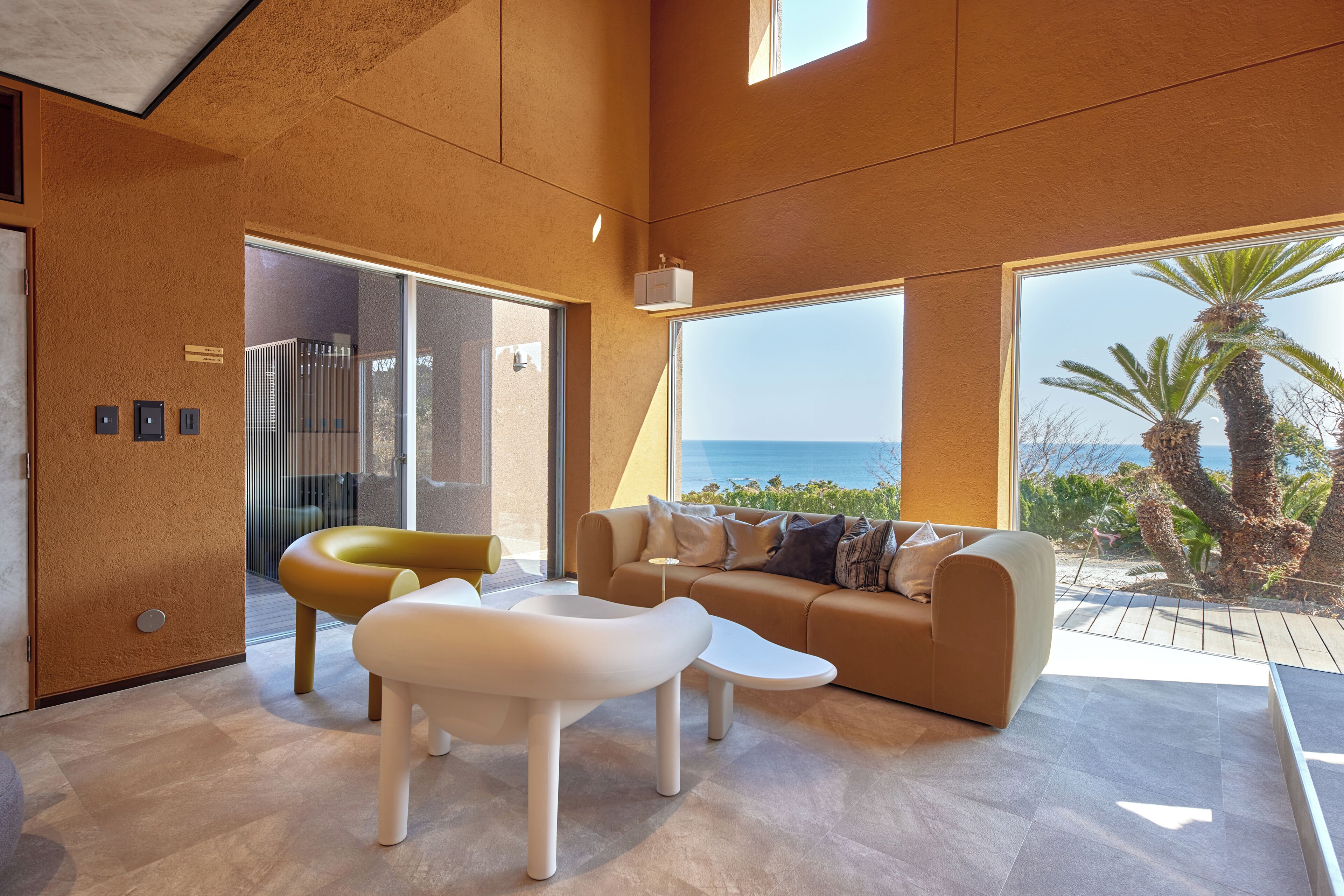 A modern living room with a sofa, two chairs, and a coffee table. There are large windows offering a view of the sea and palm trees.