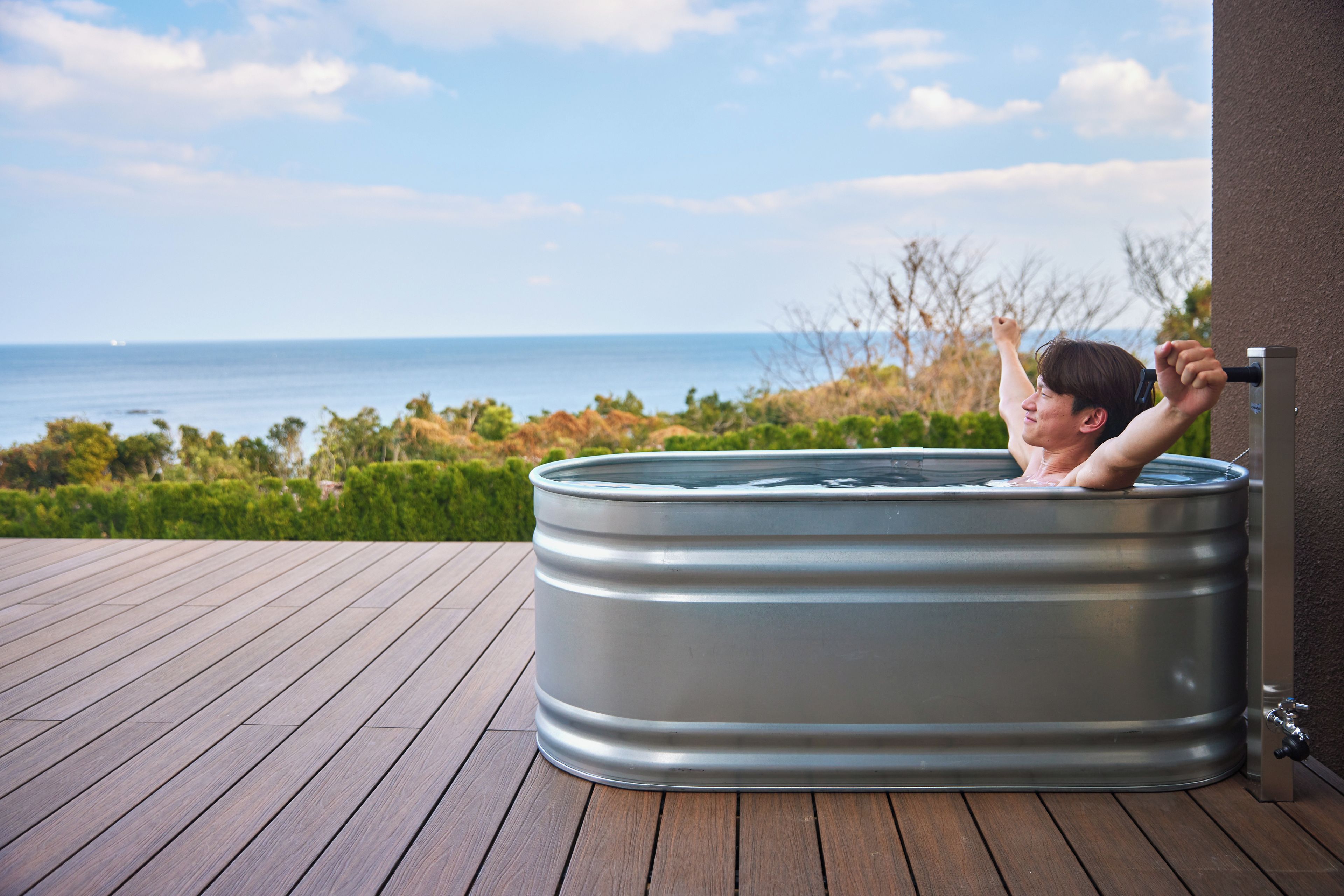A person relaxing in a hot tub on a wooden deck with a view of the ocean.