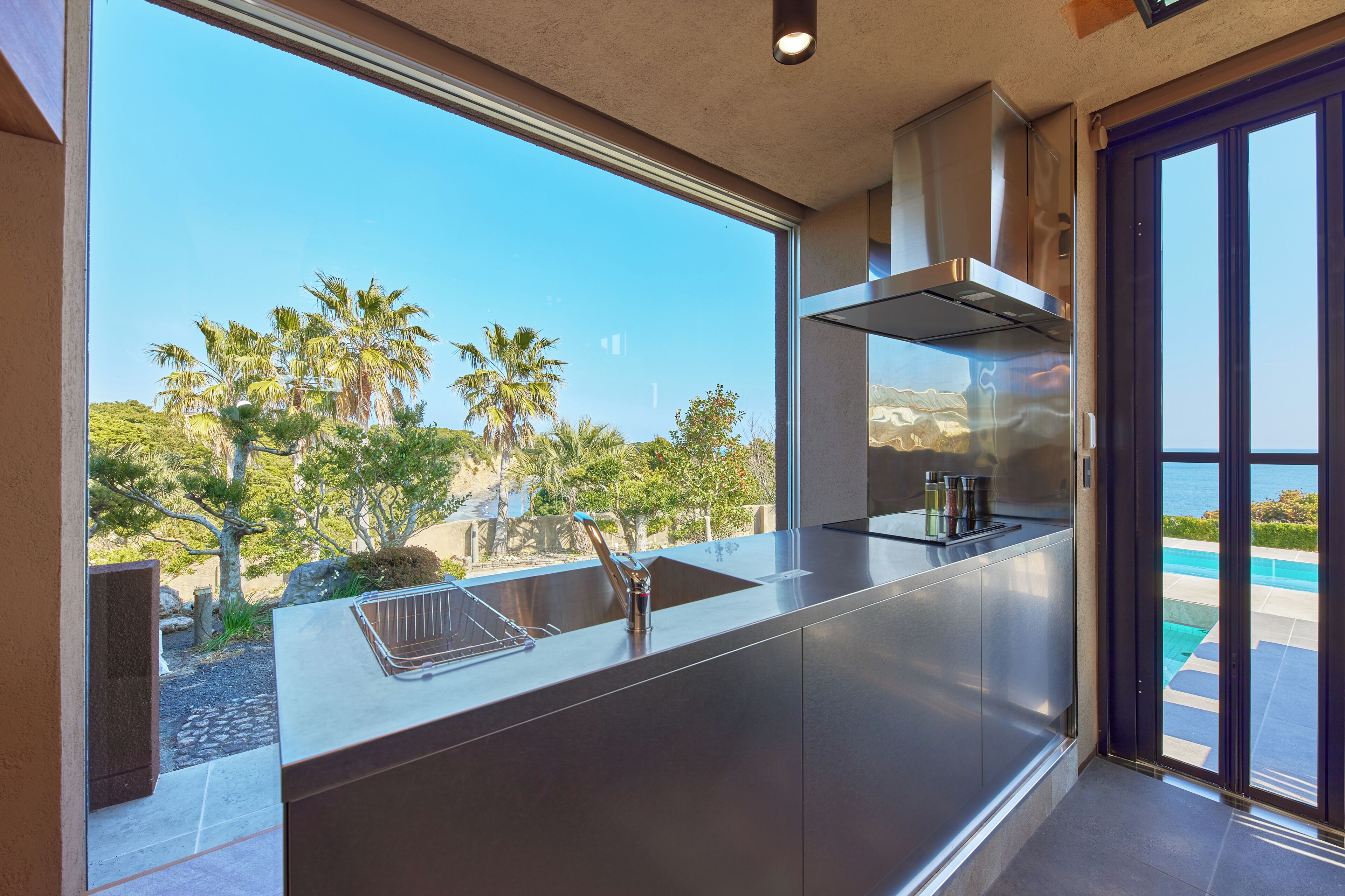 A modern kitchen with a stainless steel island, a built-in oven, and a view of a pool and palm trees.