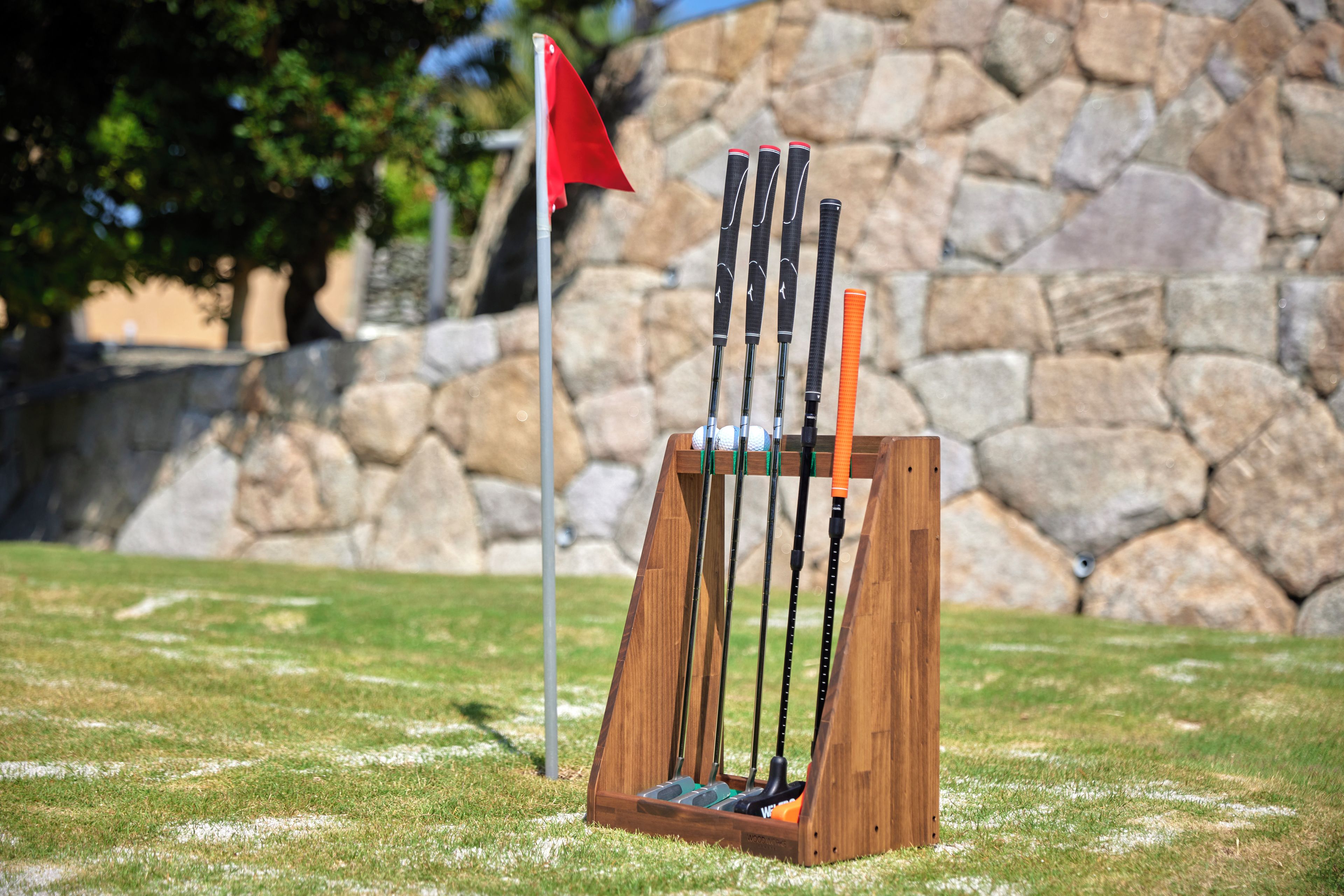 Un juego de palos de golf sobre un soporte de madera, con un muro de piedra y una bandera al fondo.