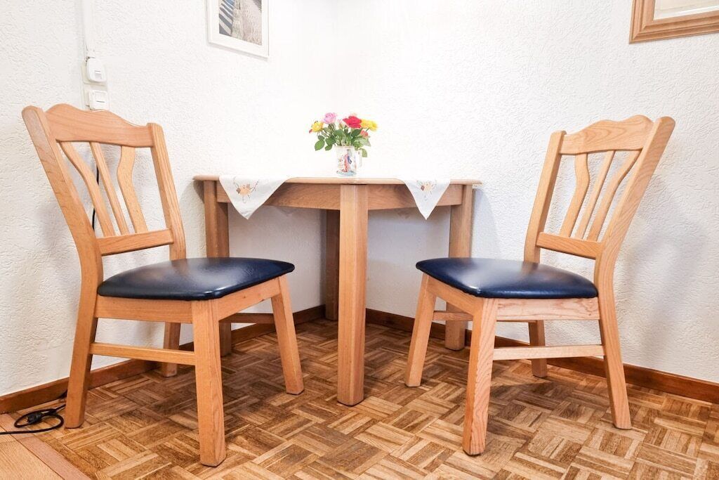 Two wooden chairs with black cushions, a small wooden table with a white cloth, and a vase with flowers on the table.