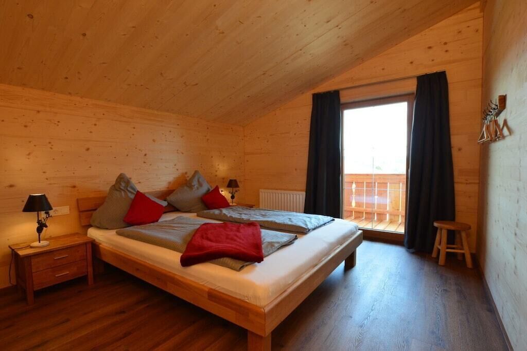 Ein Schlafzimmer einer Holzhütte mit einem Doppelbett, zwei Nachttischen, einem Beistelltisch, einem Fenster mit Vorhängen und einem hölzernen Hocker.