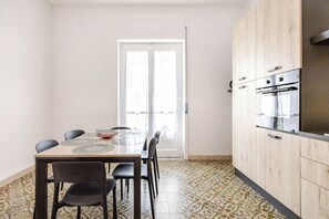 A compact kitchen with a dining area, featuring a tiled floor, a wooden cabinet with a built-in oven, and a glass-top dining table with chairs. - Mirea Domus - Salerno - Train Station/Bus/Ferry to Amalfi, 5 minutes away (Salerno)