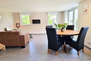 A modern living room with a sofa, a dining table with chairs, a potted plant, and a flat-screen TV mounted on the wall. - AK13 Holiday home near Domburg (Aagtekerke)