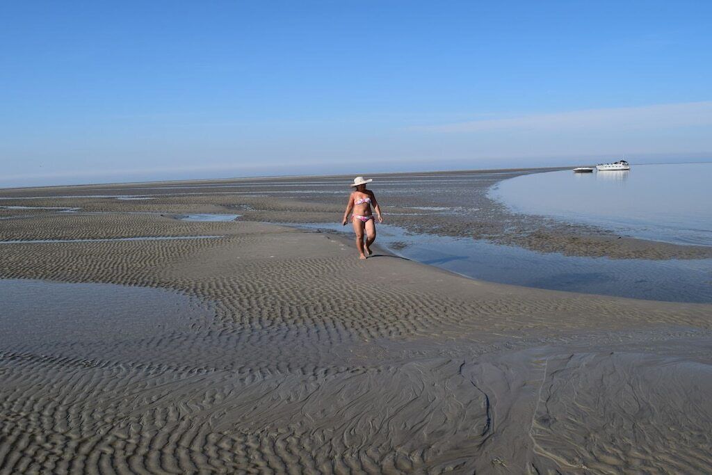 A beach with a person walking, a boat in the water, and a clear sky.