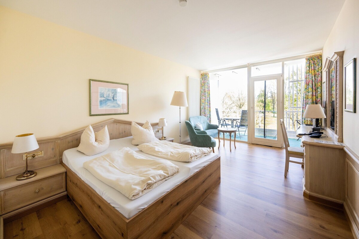 A hotel room with a wooden bed, two bedside tables with lamps, a chair, a desk, and a balcony with a view.