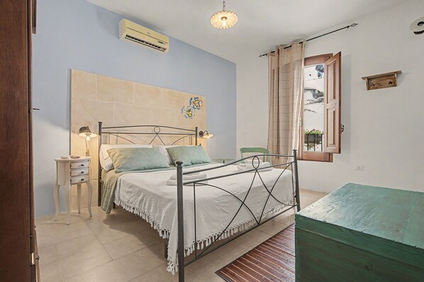 A bedroom with a bed, bedside table, lamp, air conditioning unit, and a window with curtains.