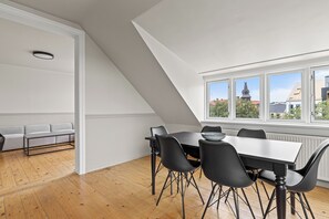 Un espace de repas moderne avec une table et des chaises, une vue sur le clocher d’une église à travers de grandes fenêtres, et un canapé gris en arrière-plan.