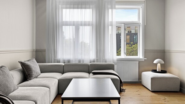 Ein modernes Wohnzimmer mit einem grauen Sofa, einem kleinen Tisch und einem Fenster mit durchscheinenden Vorhängen.