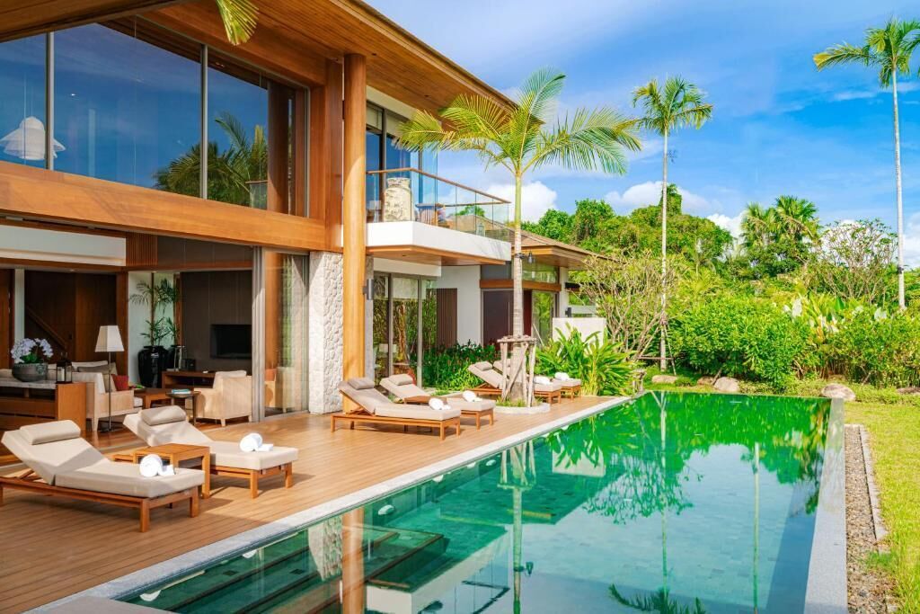 Ett modernt hus med en swimmingpool, solstolar och en balkong med utsikt över frodig grönska.
