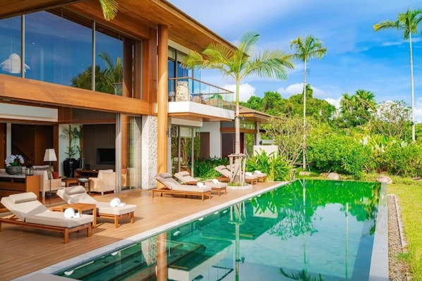A modern house with a swimming pool, lounge chairs, and a balcony overlooking lush greenery.