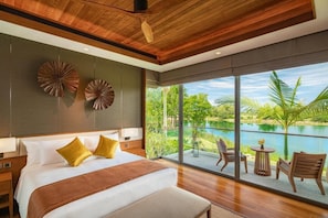 A modern hotel room with a large bed, a balcony with a view of a lake, and a dining area with chairs and a table.