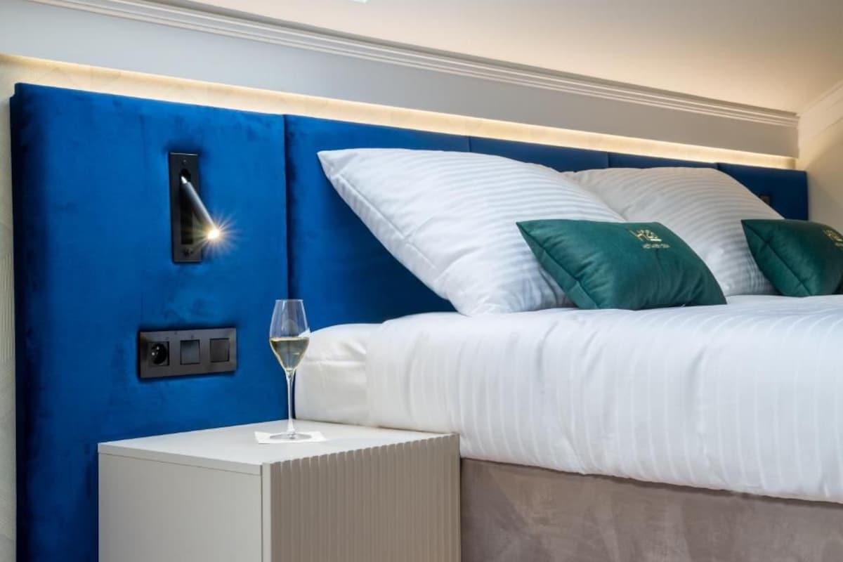 Una camera da letto con una testiera blu, biancheria bianca e un comodino con un bicchiere di vino.
