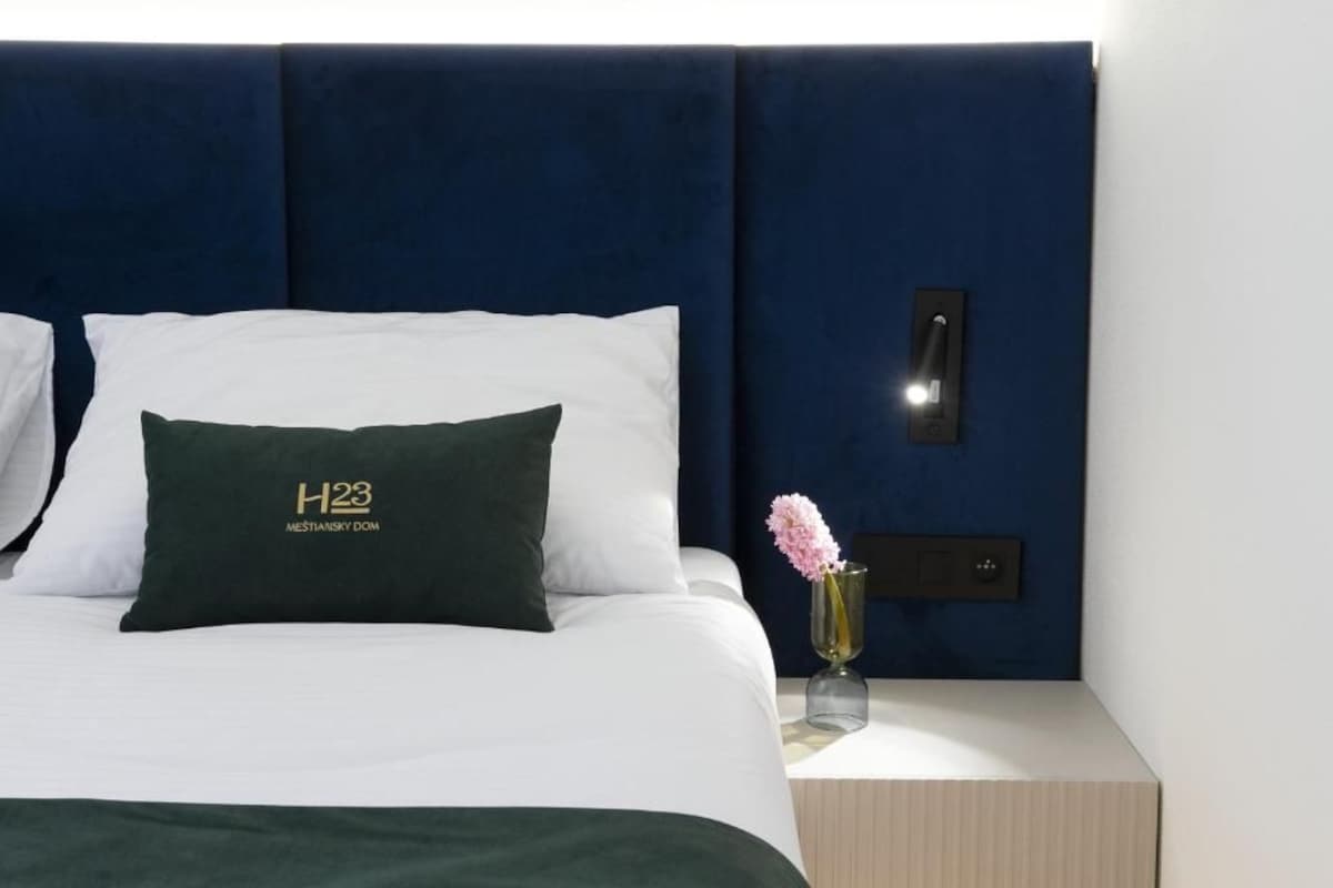 Camera d'albergo con una testata del letto blu scuro, biancheria da letto bianca, un cuscino verde con un logo, un comodino con un vaso di fiori e un pannello di controllo a parete.