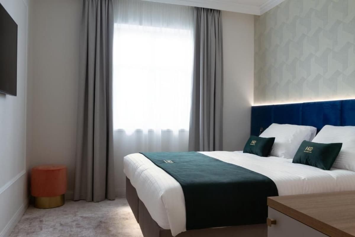Una camera d'albergo con un letto, un comodino, uno sgabello e una finestra con tende.