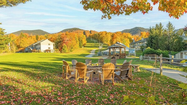 A grassy outdoor area with Adirondack chairs, a gazebo, and a view of houses and hills in the background.