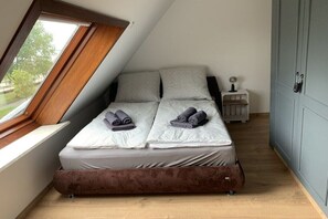 Una habitación pequeña con cama individual, techo inclinado, ventana, mesita de noche con lámpara y puerta.