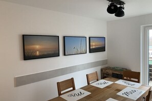 Ein Essbereich mit einem Tisch für vier Personen, drei gerahmte Bilder an der Wand und einer modernen Leuchte darüber.