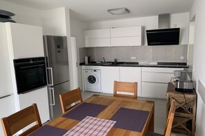 Eine kleine Küche mit Kühlschrank, Waschmaschine und Mikrowelle. Es gibt einen Esstisch mit Stühlen und einen kleinen Holznachttisch.