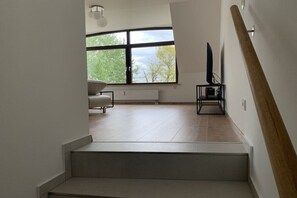 Ein modernes Interieur mit einer Treppe, einem Sofa, einem Fernseher auf einem Tisch und einem großen Fenster mit Blick auf Bäume.