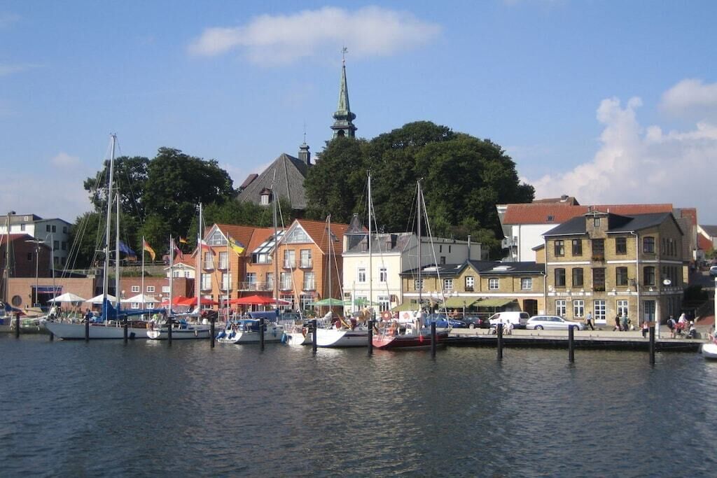 Un port de plaisance avec des bateaux amarrés, une église avec un clocher et des bâtiments aux toits rouges.