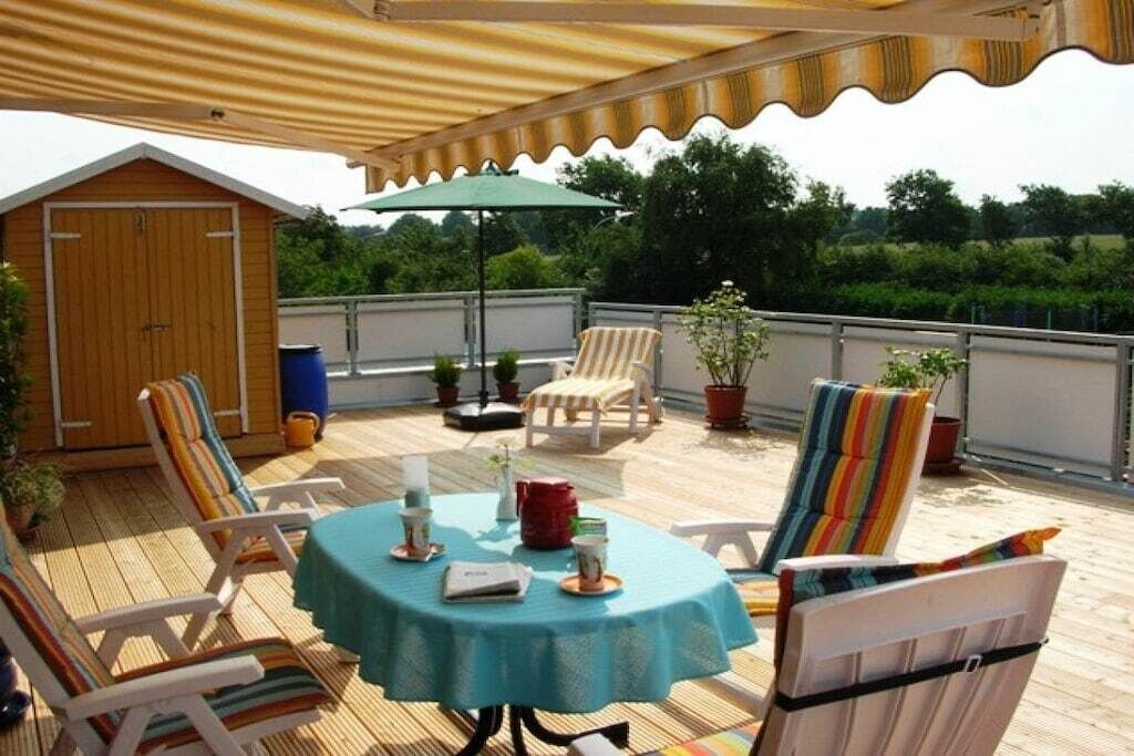 Ein überdachter Sitzbereich im Freien mit Tisch und Stühlen, ein Holzgebäude und Kübelpflanzen.