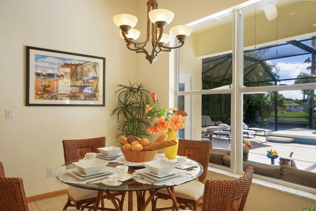 Un comedor con una mesa redonda de vidrio, sillas de mimbre, una lámpara de araña, una planta en maceta y un ventanal con vista a muebles de exterior.