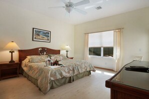 Ein Schlafzimmer mit einem Bett, zwei Nachttischen, einem Deckenventilator, einem Fenster mit Vorhängen und einem Schreibtisch mit Stuhl.