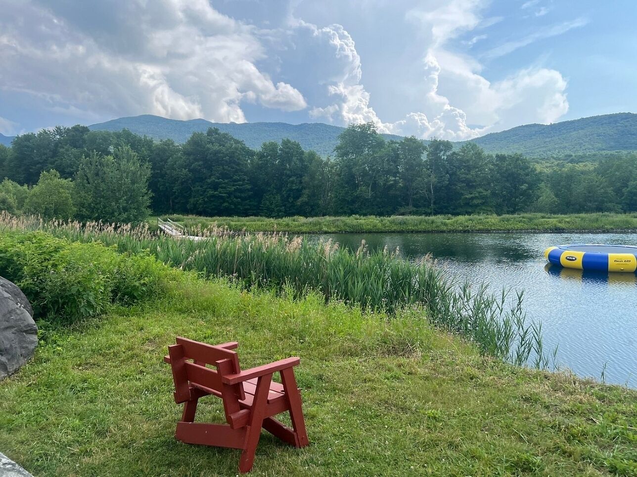 A lakeside view with a red bench, a floating dock, and a forested hill in the background.