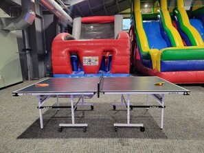 Två bordtennisbord i ett inomhusutrymme med en uppblåsbar rutschkana och en röd lekställning i bakgrunden.