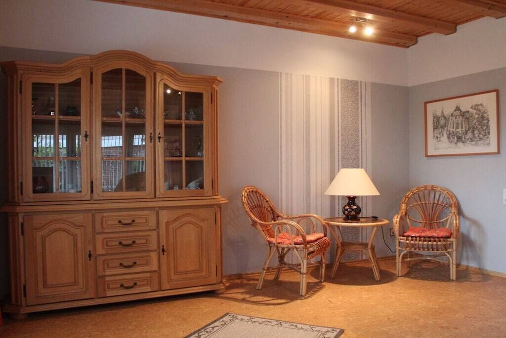 Ett rum med ett träskåp, två fåtöljer i rotting, ett litet bord med en lampa och en inramad tavla på väggen.