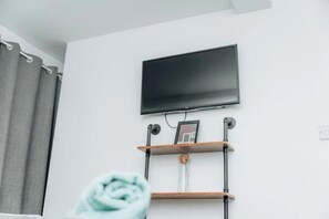 Une pièce avec un téléviseur à écran plat fixé au mur, une étagère en bois avec un cadre photo et un rideau.