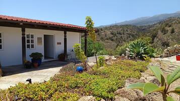 A building with a red roof, a garden with various plants, and a mountainous landscape in the background.