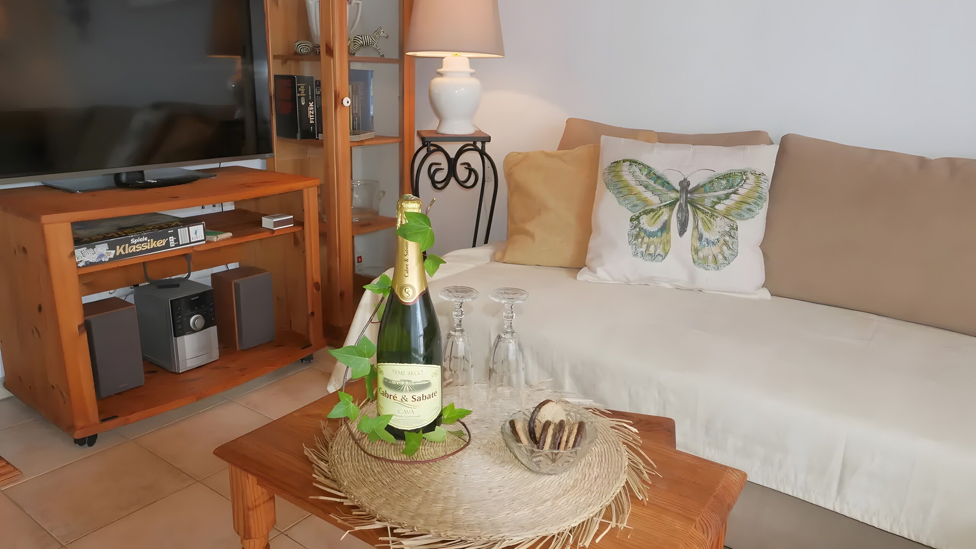 Ein Wohnzimmer mit einer Holz-Unterhaltungsecke, einem Fernseher, einem Sofa mit einem Schmetterlingskissen und einem Couchtisch mit einer Champagnerflasche und Gläsern.